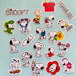 May include: A selection of Snoopy stickers in different poses and scenes. The stickers depict Snoopy with Charlie Brown, in a dinosaur costume, on a skateboard, and other designs. The words "Snoopy" and "Peanuts" are also visible.