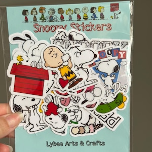May include: A pack of Snoopy stickers in a clear plastic sleeve. The stickers depict Snoopy, Charlie Brown, and other Peanuts characters. The words "Snoopy Stickers" and "Lybee Arts & Crafts" are printed on the backing card.
