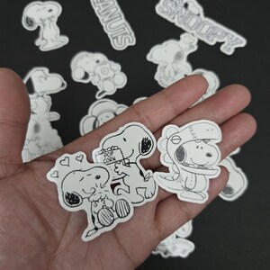 May include: A selection of black and white Snoopy stickers, with the words "PEANUTS" and "SNOOPY" in white. The stickers depict Snoopy in different poses, including holding popcorn and wearing a hat. A fun collection.