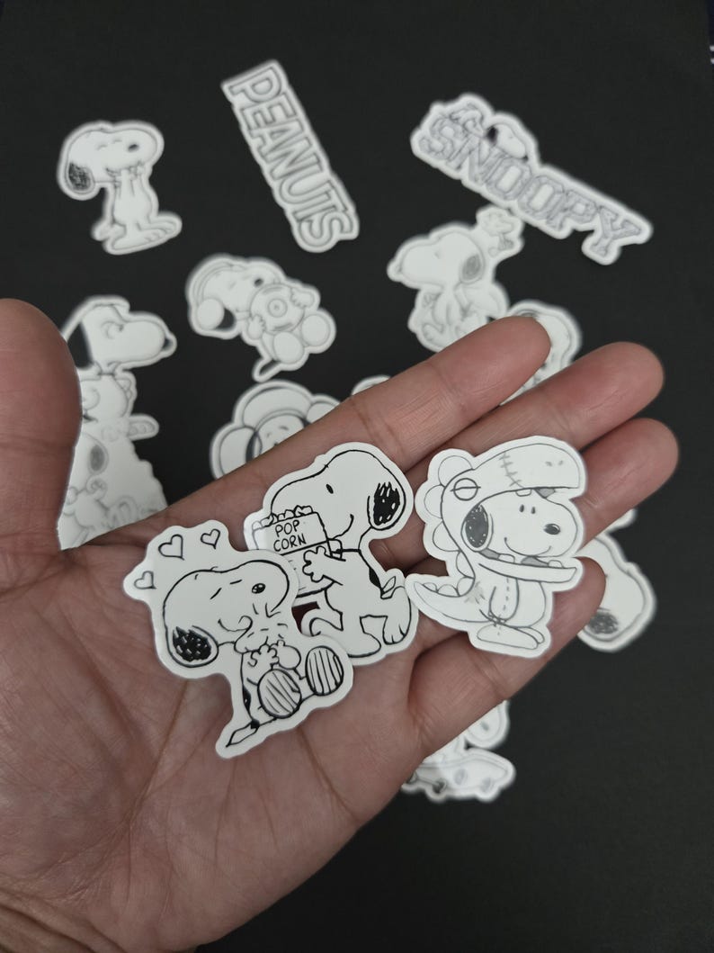 May include: A selection of black and white Snoopy stickers, with the words "PEANUTS" and "SNOOPY" in white. The stickers depict Snoopy in different poses, including holding popcorn and wearing a hat. A fun collection.