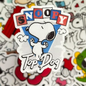 May include: A Snoopy sticker featuring the word "SNOOPY" in red, blue, and white blocks above a white Snoopy character. The words "Top Dog" are written below Snoopy in a cursive font. The background is a light blue triangle.