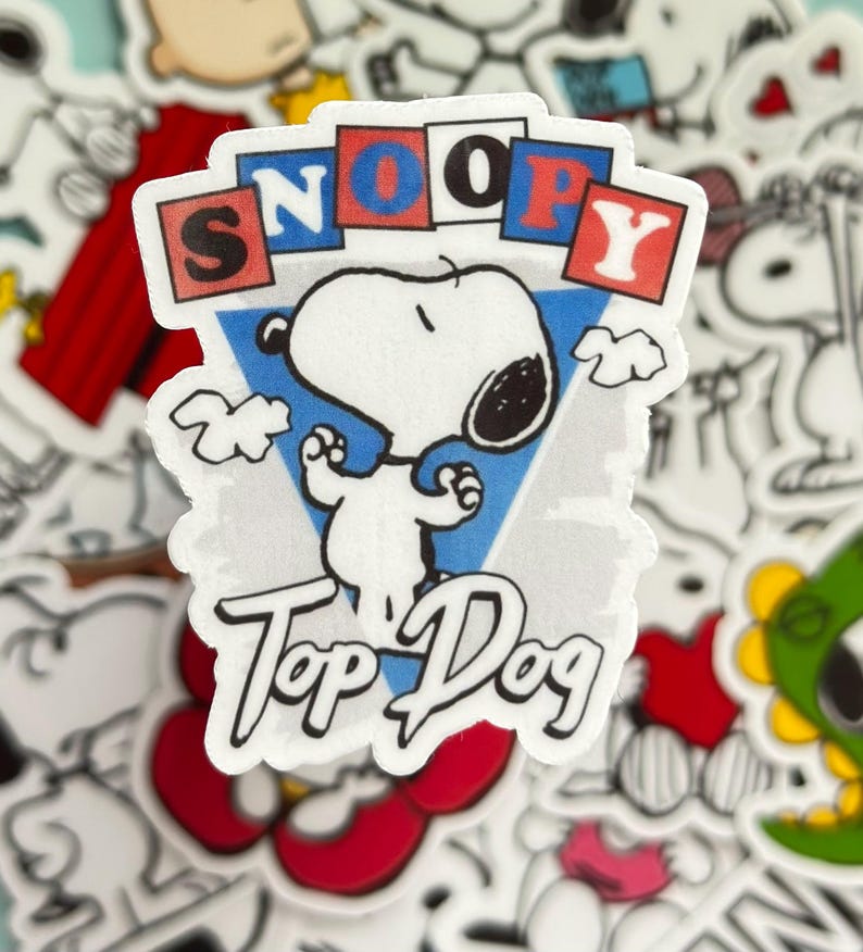 May include: A Snoopy sticker featuring the word "SNOOPY" in red, blue, and white blocks above a white Snoopy character. The words "Top Dog" are written below Snoopy in a cursive font. The background is a light blue triangle.