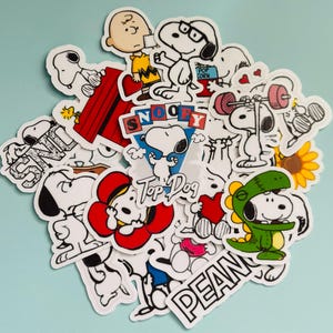 May include: A selection of Snoopy stickers in different poses and costumes. The stickers depict Snoopy with Charlie Brown, Snoopy lifting weights, and Snoopy in a dinosaur outfit. The stickers are white with black outlines and colourful details.