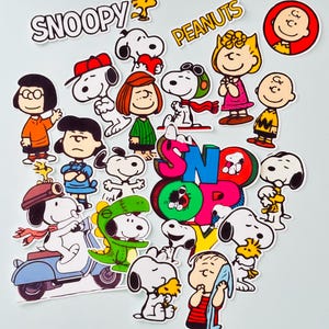 May include: A selection of colourful Snoopy and Peanuts character stickers. The stickers feature Snoopy in various poses, along with Charlie Brown, Lucy, Linus, and other characters. The words "Snoopy" and "Peanuts" are also displayed.