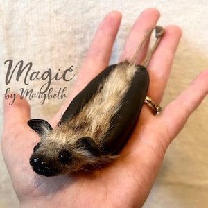May include: A handcrafted bat decoration with a black body, tan fur, and small black ears. The bat has a string for hanging and the text "Magic by Marybeth" is visible in the image.