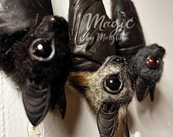 May include: Close-up of three handcrafted bat figures with detailed fur and leather wings. The bats have large, glossy eyes and are in varying shades of black, gray, and brown. The text "Magic by Marybeth" is visible.