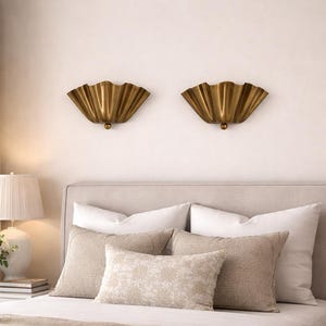 May include: Two gold-colored wall sconces with a fan-like design are mounted above a bed with white and beige pillows. The sconces have a textured surface and a small, round detail at the bottom. The room has a neutral color palette.