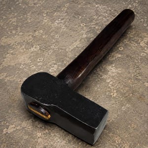 Op de afbeelding: Een robuuste hamer met een massieve zwarte metalen kop met een gladde, gepolijste afwerking en een donkerbruine houten steel. De kop is stevig aan de steel bevestigd met een zichtbare houten wig, wat een strak en functioneel ontwerp laat zien.