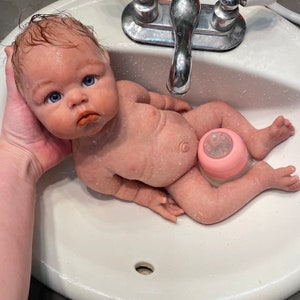 May include: A realistic baby doll with blue eyes and wet hair is held in a white sink. The doll has a pink bottle attached to its leg. The doll's skin has a realistic texture and is wet.