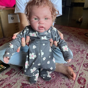 May include: A realistic baby doll with fair skin, green eyes, and curly red hair. The doll is wearing a gray onesie with a white and pink floral pattern. The onesie has a zipper down the front.