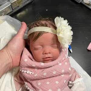 May include: A lifelike baby doll with closed eyes, wearing a pink and white polka dot blanket and a matching outfit. A white flower and a cream-colored headband adorn the doll's head. The doll is being held by a person.