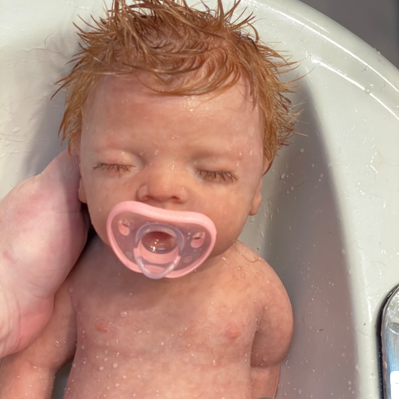 May include: A reborn doll with wet, reddish-blonde hair and closed eyes. The doll has a pink pacifier in its mouth and is in a white bathtub. Water droplets cover the doll's skin.