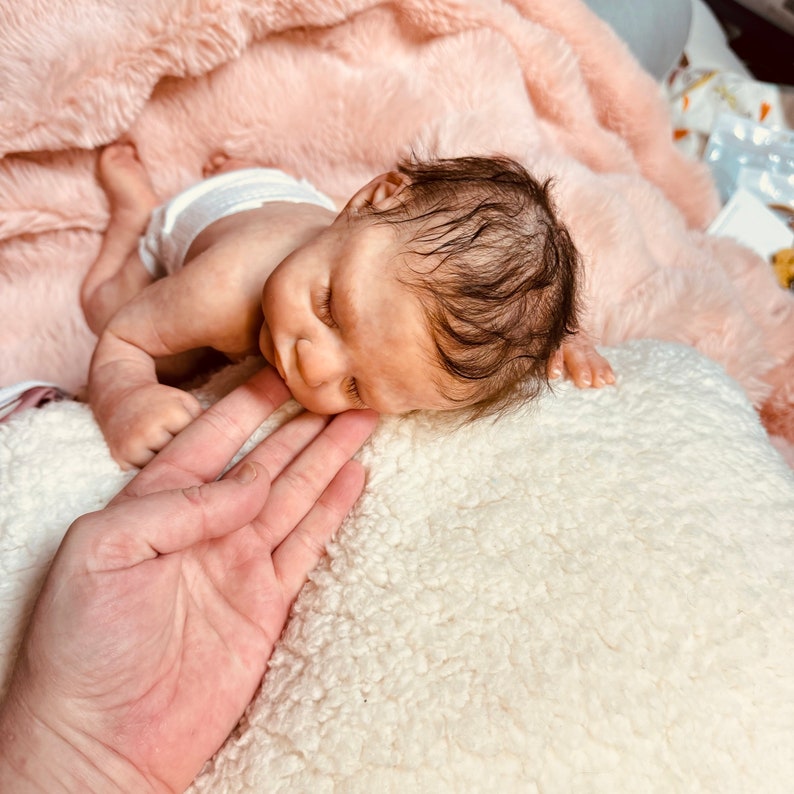May include: A realistic baby doll with detailed skin tones and dark hair, wearing a diaper. The doll is lying on a white, fluffy surface, with a pink blanket in the background. A hand is gently touching the doll's face.