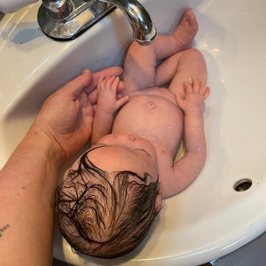 May include: A baby is bathed in a white porcelain sink under a chrome faucet. The baby is wet, with dark hair and is being held by a person's hand. The baby's skin is light-toned, and water droplets are visible on the baby's body.