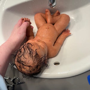 May include: A realistic baby doll with dark hair lies in a white sink. The doll's skin has a lifelike tone, and it appears to be wet. A hand gently holds the doll's foot. The sink is clean and has a silver faucet.