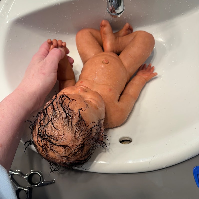May include: A realistic baby doll with dark hair lies in a white sink. The doll's skin has a lifelike tone, and it appears to be wet. A hand gently holds the doll's foot. The sink is clean and has a silver faucet.