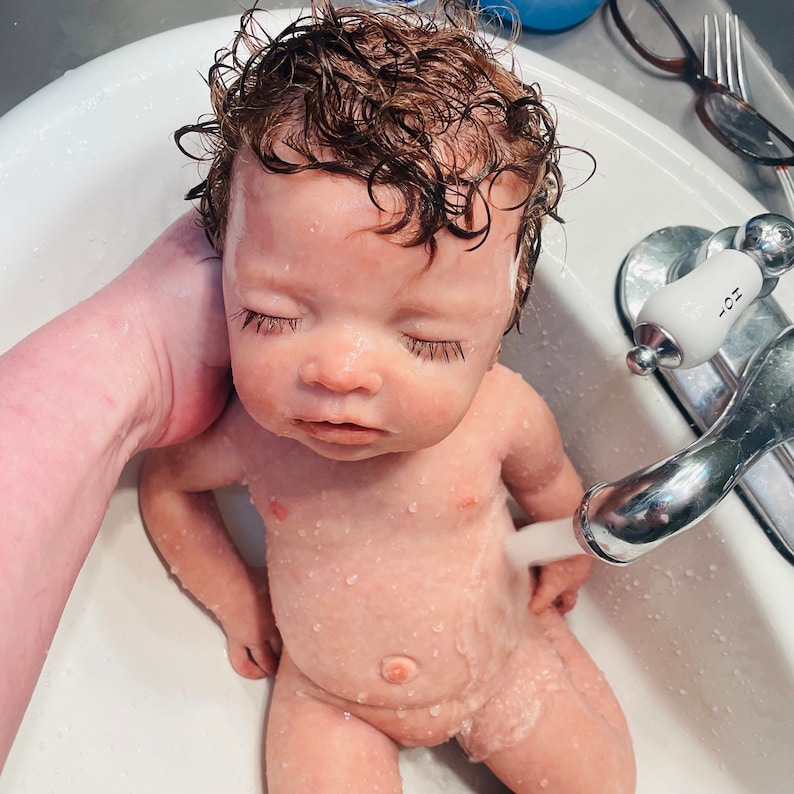 May include: A baby doll with closed eyes is being bathed in a white sink. The doll has wet, curly brown hair and water droplets on its skin. A hand is holding the doll, and a faucet is running water over it.