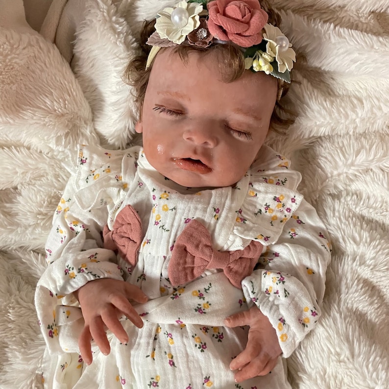 May include: A lifelike baby doll wearing a white floral print dress with pink bows. The doll has closed eyes, light skin, and a floral headband with pink and cream-colored flowers. The doll is lying on a fluffy white surface.