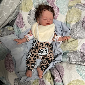 May include: A realistic baby doll with closed eyes, wearing a cream-colored bib and patterned pants. The pants feature a cow skull design on a tan background with black spots. The doll is lying on a bed with a floral patterned comforter.