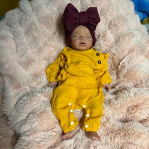 May include: A baby doll wearing a mustard yellow jumpsuit with white snaps and a burgundy bow headband. The doll is lying on a pink, fluffy blanket. The jumpsuit has long sleeves and decorative wooden buttons.