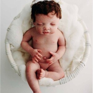 May include: A newborn baby with dark brown hair and a white floral headband sits in a round, woven basket lined with fluffy white fabric. The baby is nude, with closed eyes, and is positioned in a seated pose.