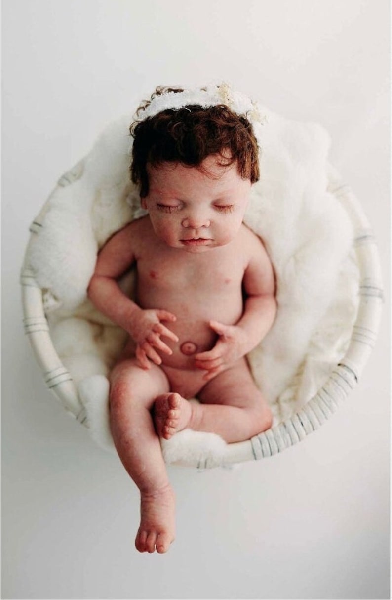 May include: A newborn baby with dark brown hair and a white floral headband sits in a round, woven basket lined with fluffy white fabric. The baby is nude, with closed eyes, and is positioned in a seated pose.