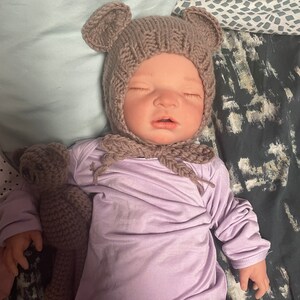 May include: A baby wearing a knitted brown bear bonnet with matching bear ears and a lavender long-sleeved onesie. A small crocheted teddy bear is next to the baby. The baby is lying down with its eyes closed.
