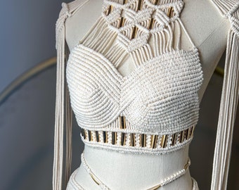 May include: A cream-coloured macrame two-piece set featuring a halter-style crop top with intricate knotting, gold-toned cylindrical accents, and long fringe details. The matching skirt includes a braided waist tie with gold-toned hardware.
