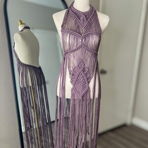 May include: A lavender macrame halter top with intricate knotting and fringe detailing. The handmade top is displayed on a mannequin, showcasing its open back and decorative patterns. The top is a unique piece of bohemian fashion.