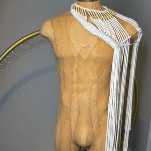 Peut inclure: Un collier en macram&eacute; blanc avec des accents dor&eacute;s. Le collier est drap&eacute; sur un torse de mannequin beige.