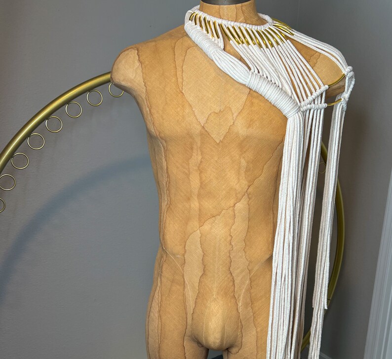 Peut inclure: Un collier en macram&eacute; blanc avec des accents dor&eacute;s. Le collier est drap&eacute; sur un torse de mannequin beige.