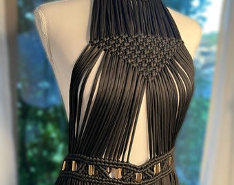 May include: A black macrame halter top featuring intricate knotted patterns at the neckline and a waist section accented with metallic gold cylindrical beads. The design consists of long, vertical cord strands creating a fringe effect throughout.