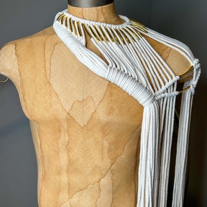 Peut inclure: Un collier en macram&eacute; blanc avec des accents dor&eacute;s. Le collier est fait d'un cordon &eacute;pais et a un design g&eacute;om&eacute;trique. Il est port&eacute; sur un mannequin.