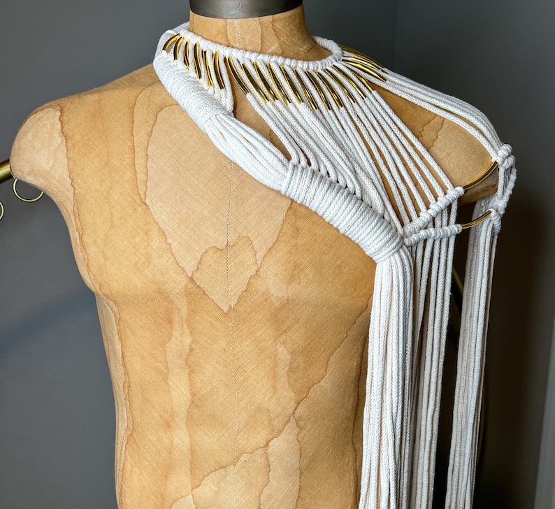 Peut inclure: Un collier en macram&eacute; blanc avec des accents dor&eacute;s. Le collier est fait d'un cordon &eacute;pais et a un design g&eacute;om&eacute;trique. Il est port&eacute; sur un mannequin.