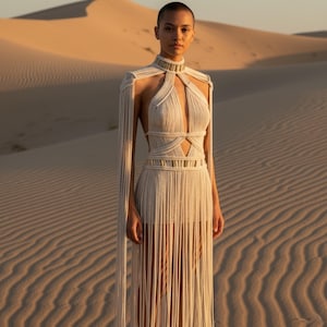 May include: A cream-colored macrame dress featuring a high neck, a deep cutout bodice, and long, flowing fringe sleeves. The garment is adorned with horizontal bands of metallic gold accents at the neck and waist, creating a structured look.