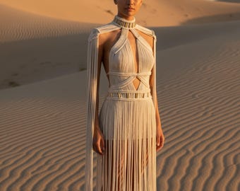 Puede incluir: Un vestido de macramé color crema con cuello alto, un corpiño con aberturas profundas y mangas largas de flecos fluidos. La prenda está adornada con bandas horizontales con detalles en oro metálico en el cuello y la cintura.