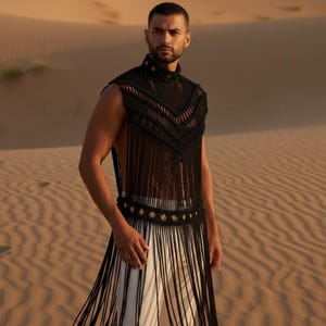 May include: A black macrame top with fringe detail and gold accents. The top is paired with white pants. The model is standing in a desert setting with sand dunes in the background. The top has a high collar and decorative buttons.