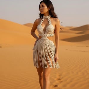 May include: A cream-colored macrame dress with a halter top, cut-out midriff, and fringed skirt. The dress is worn in a desert setting with sand dunes in the background. The dress has a choker-style neckline and intricate woven details.