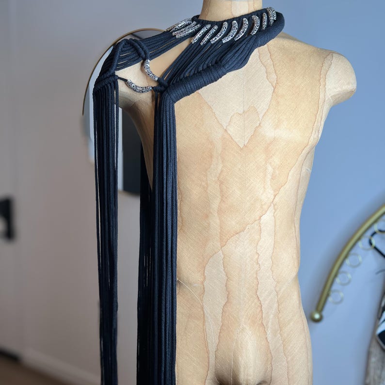 Peut inclure: Un collier ras de cou en tissu noir avec des embellissements argent&eacute;s et de longues franges. Le collier est drap&eacute; sur l'&eacute;paule d'un mannequin.
