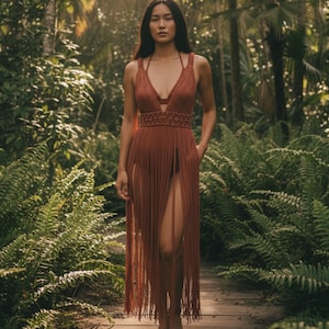 Könnte beinhalten: Ein rostfarbenes Kleid mit Fransen, tiefem Ausschnitt und hohen Schlitzen. Das Kleid hat einen gewebten Gürtel und dünne Träger. Das Model steht auf einem Holzsteg, umgeben von üppigem, grünem Laub.