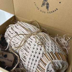 K&ouml;nnte beinhalten: Ein beigefarbener Makramee-Wandbehang mit goldenen Akzenten ist in einem braunen Karton verpackt, auf dem der Text "ENTRE DOS TIERRAS" aufgedruckt ist. Der Makramee ist mit Schnur gebunden und von zerkleinertem Papier umgeben.