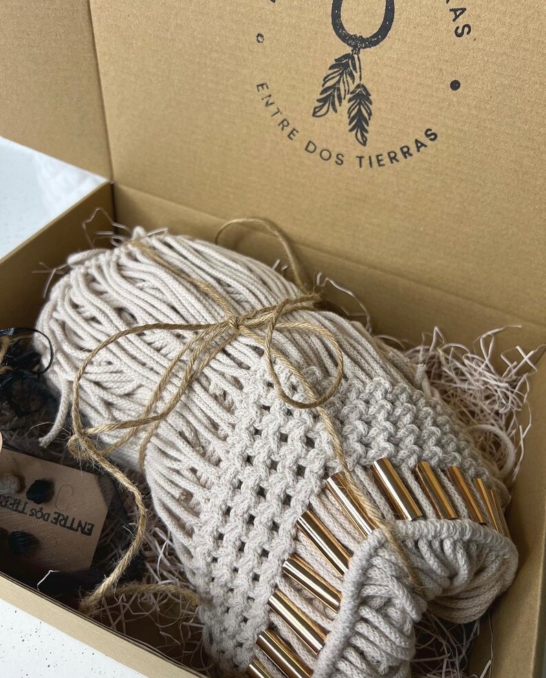 K&ouml;nnte beinhalten: Ein beigefarbener Makramee-Wandbehang mit goldenen Akzenten ist in einem braunen Karton verpackt, auf dem der Text "ENTRE DOS TIERRAS" aufgedruckt ist. Der Makramee ist mit Schnur gebunden und von zerkleinertem Papier umgeben.