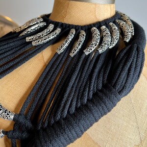 Peut inclure: Un collier en cordon de coton noir avec des accents en m&eacute;tal argent&eacute;. Le collier est compos&eacute; de nombreuses m&egrave;ches de cordon qui sont tiss&eacute;es ensemble pour cr&eacute;er un aspect &eacute;pais et textur&eacute;. Les accents en m&eacute;tal argent&eacute; sont en forme de petites boucles orn&eacute;es.