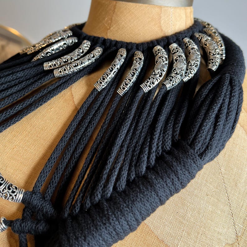 Peut inclure: Un collier en cordon de coton noir avec des accents en m&eacute;tal argent&eacute;. Le collier est compos&eacute; de nombreuses m&egrave;ches de cordon qui sont tiss&eacute;es ensemble pour cr&eacute;er un aspect &eacute;pais et textur&eacute;. Les accents en m&eacute;tal argent&eacute; sont en forme de petites boucles orn&eacute;es.