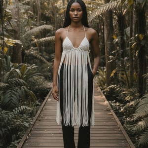 May include: A cream-colored, halter-style top with a fringed design. The top is paired with black pants. The image is set in a lush, green forest with a wooden walkway.