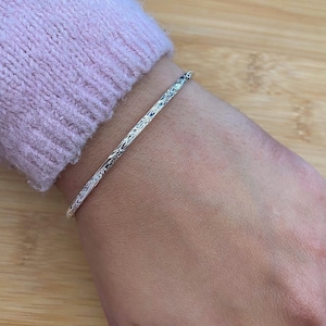 May include: A silver bangle bracelet with a textured surface. The bracelet is thin and appears to be made of metal. The bracelet is worn on a wrist, with a light pink sweater in the background.