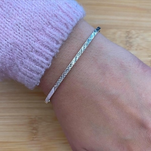 May include: A silver bracelet with a textured, geometric pattern. The bracelet is thin and sits on a wrist, with a light purple sweater in the background. The bracelet is a simple, elegant piece of jewelry.