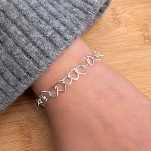 Könnte beinhalten: Ein silberfarbenes Armband mit einem kettenartigen Design aus miteinander verbundenen Herzformen. Das Armband wird am Handgelenk getragen, im Hintergrund ist ein grauer Ärmel zu sehen. Das Armband besteht aus glänzendem Metall.