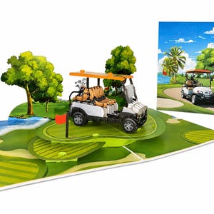 Könnte beinhalten: Eine dreidimensionale Pop-up-Grußkarte mit einem weißen Golfcart mit orangefarbenem Dach, hellbraunen Sitzen und Golfschlägern im Heck. Die Karte zeigt eine grüne Golfplatzlandschaft mit Bäumen, einem Wasserhindernis und einer roten Fahne.