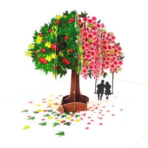 Op de afbeelding: Een 3D pop-up kaart met een boom met twee helften: één met groene bladeren en rode appels, de andere met roze bloesems. Een silhouet van een stel op een schommel hangt aan de bloeiende kant. Verspreide bladeren en bloemblaadjes.